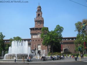 a large brick building with a clock tower with Sforza Castle in the background