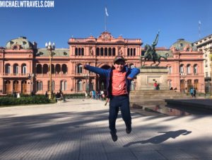 a boy jumping in front of a pink building with Casa Rosada in the background