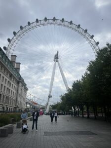 A large Ferris wheel, known as the London Eye, is prominently featured against a cloudy sky. Below, people are walking along a paved path lined with trees on one side and a historic building on the other. A person is pushing a stroller, and others are casually strolling or standing. The scene conveys a typical day in a bustling urban area.