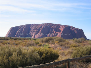 a large red rock in the desert with Uluru in the background
