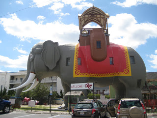 a large elephant statue in a parking lot with Lucy the Elephant in the background