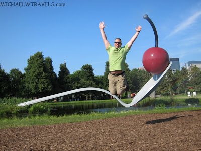 a man jumping in the air with a spoon and a cherry sculpture with Minneapolis Sculpture Garden in the background
