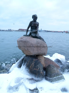 a statue of a woman sitting on a rock with The Little Mermaid in the background