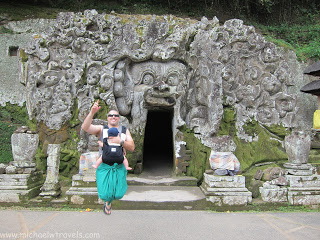 a man and child running in front of a carved stone structure with Goa Gajah in the background